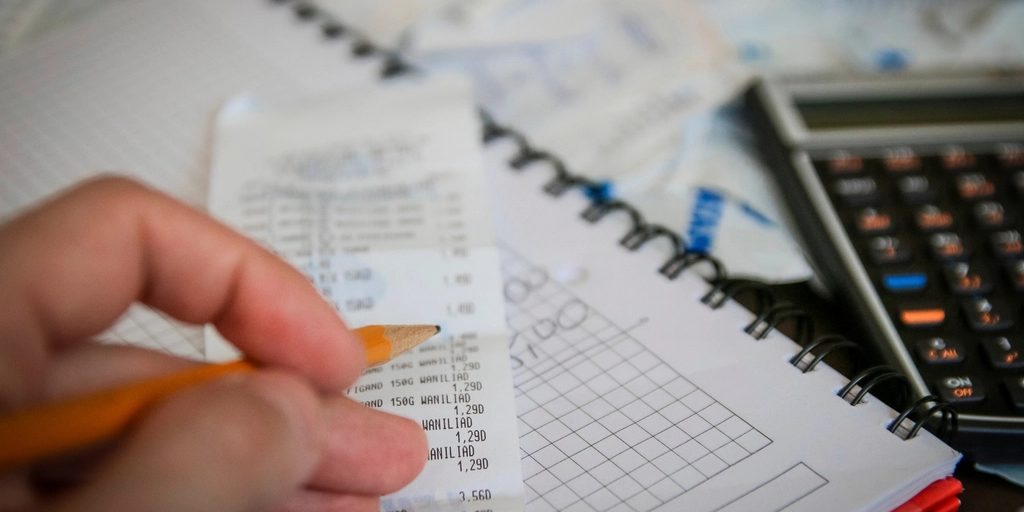 A hand writing figures on a pad beside a calculator
